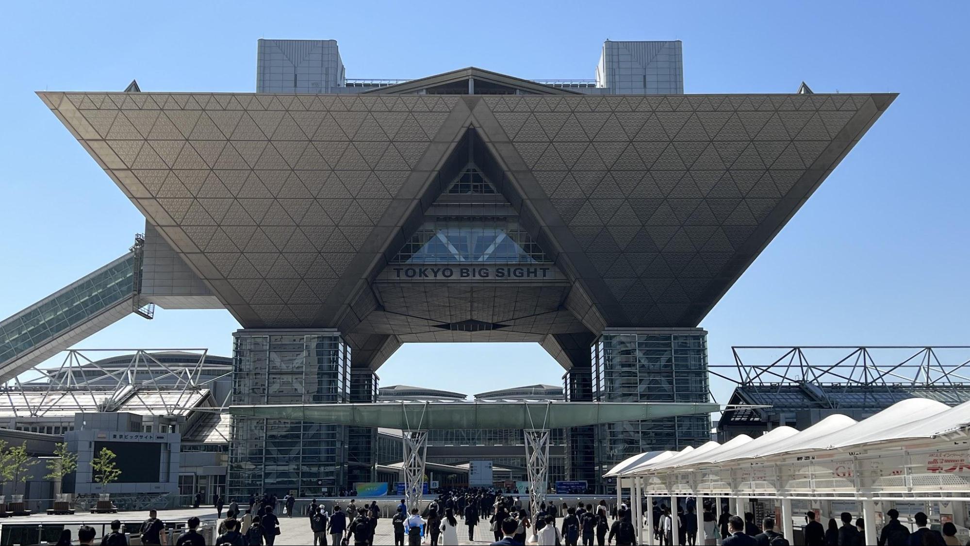 晴天の中にそびえる東京ビッグサイトと、会場に向かう人々の後ろ姿